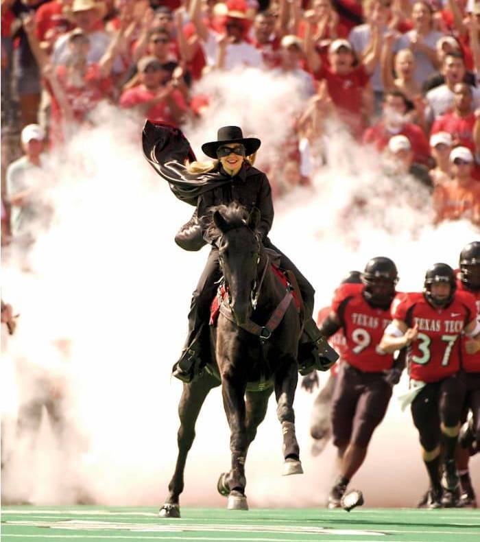The Masked Rider emerges from a puff of smoke and leads the TTU football team onto the field. A crowd of fans cheers in the background.