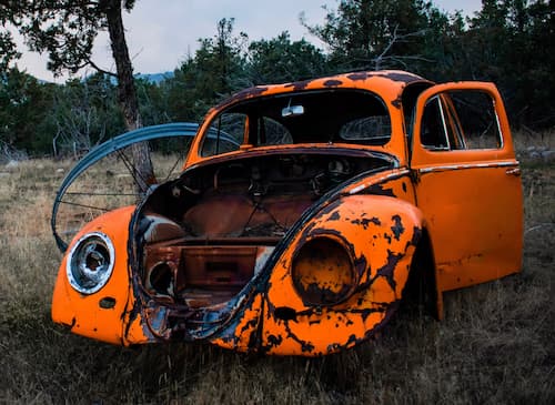 A rusty Volkswagen Beetle sits in a field. The car is missing its windshield, passenger windows, and a headlight.