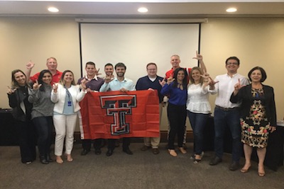 Dr. Justin Louder, Jeff Oldham and Jared Lay stand with Instructors and Coordinators as they show the guns up symbol and hold a Texas Tech flag. 