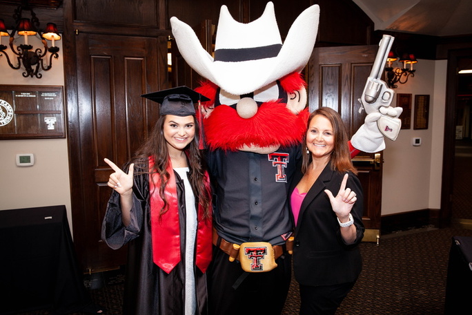 Presley Duyck stands to the right of Raider Red performing the guns up symbol while her mother stands to the right of Raider Red also performing the guns up symbol