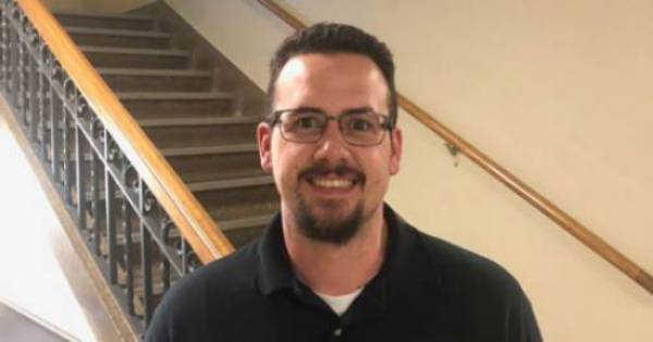 Jeremy Hogan stands in front of a staircase smiling while facing the camera 