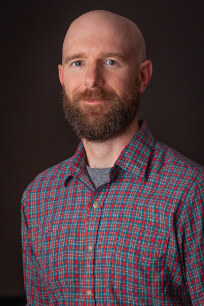 A headshot of Kyle Dawson standing against a black background and wearing a blue and red stripped shirt.