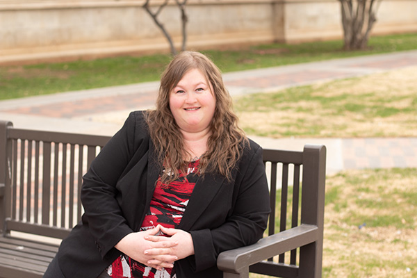 Crystal Green sits on a metal bench and leans on the arm rest while smiling and wearing a black blazer with a red and black dress underneath