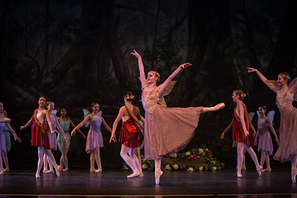 Emma Gray poses on-stage standing on one foot while surrounded by other dancers standing in front of a dark backdrop