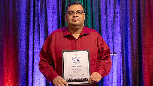 Israel Galvan stands in front of curtain that is illuminated with various colors and holds his award at waist level while looking at the camera.