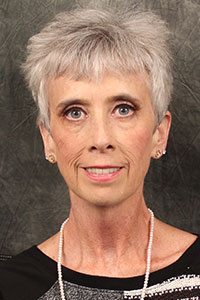 Jeanye Wester looks forward while smiling and standing in front of a dark-gray background and wearing earrings, a pearl necklace, and a black and white shirt.