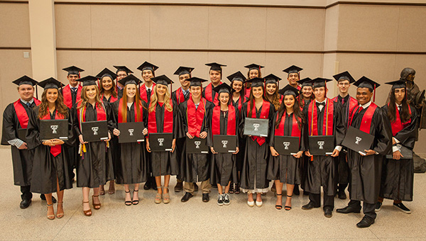A group of students face the camera while standing alongside one another and holding their individual diplomas in their hands.