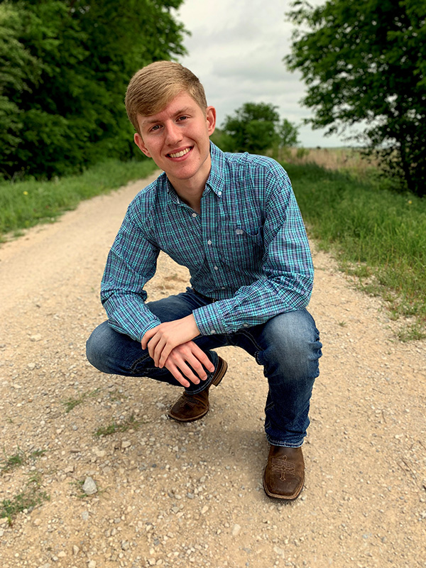 Trey Terry crouches outside on a dirt road while smiling and wearing a light-blue plaid shirt and jeans.