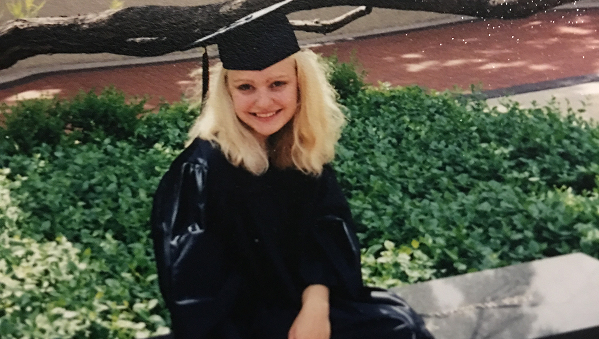 Laina wears a traditional graduation cap and gown in black and sits outside on a flat bench with a tree and greenery in the background
