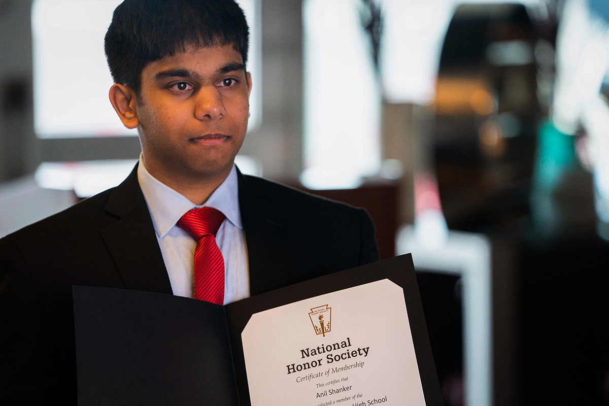 Anil Shanker A young man wearing a black suit and red tie holds a National Honor Society certificate while staring off camera