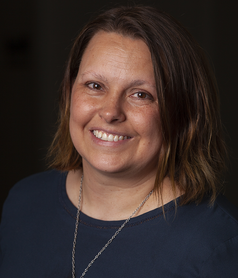 A headshot of Karen Busby while wearing a necklace and a dark blue shirt