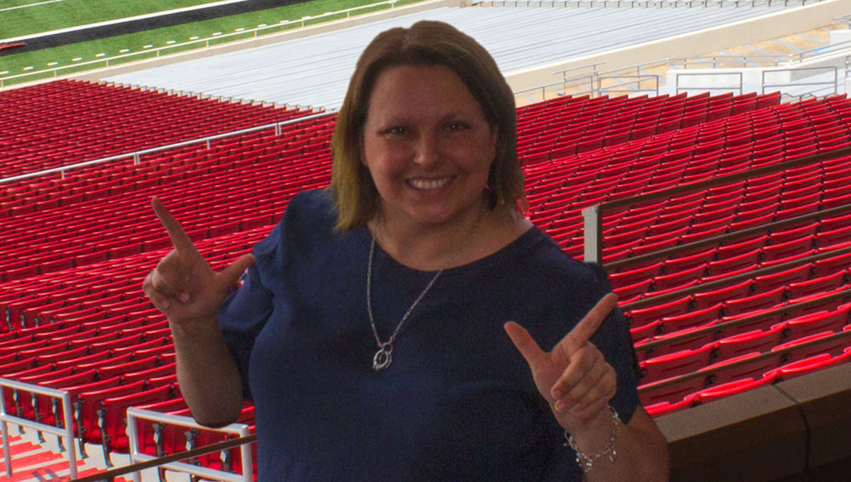 A woman wearing a blue shirt and a necklace stands above rows of red bleachers and displays the guns up symbol on both hands while smiling.