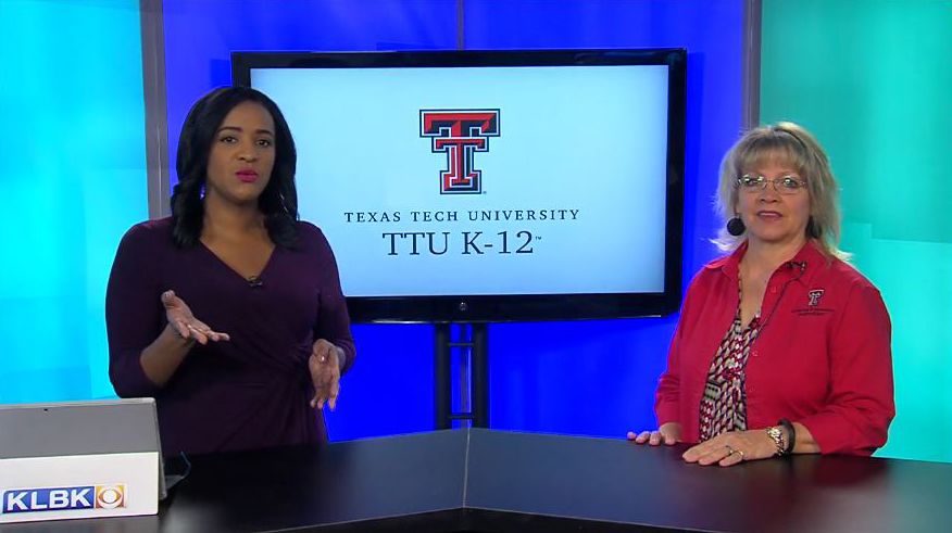 Two women stand side-by-side behind a desk facing the camera with a television behind them displaying the ttu k-12 logo 