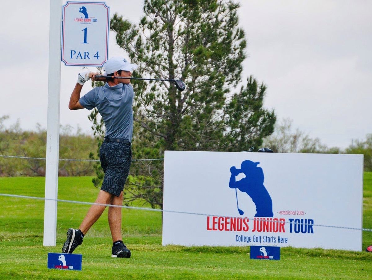 Mikey poses outside wearing a white cap and holding a golf driver while standing next to a Legends Junior Tour sign depicting a silhouette performing the same pose
