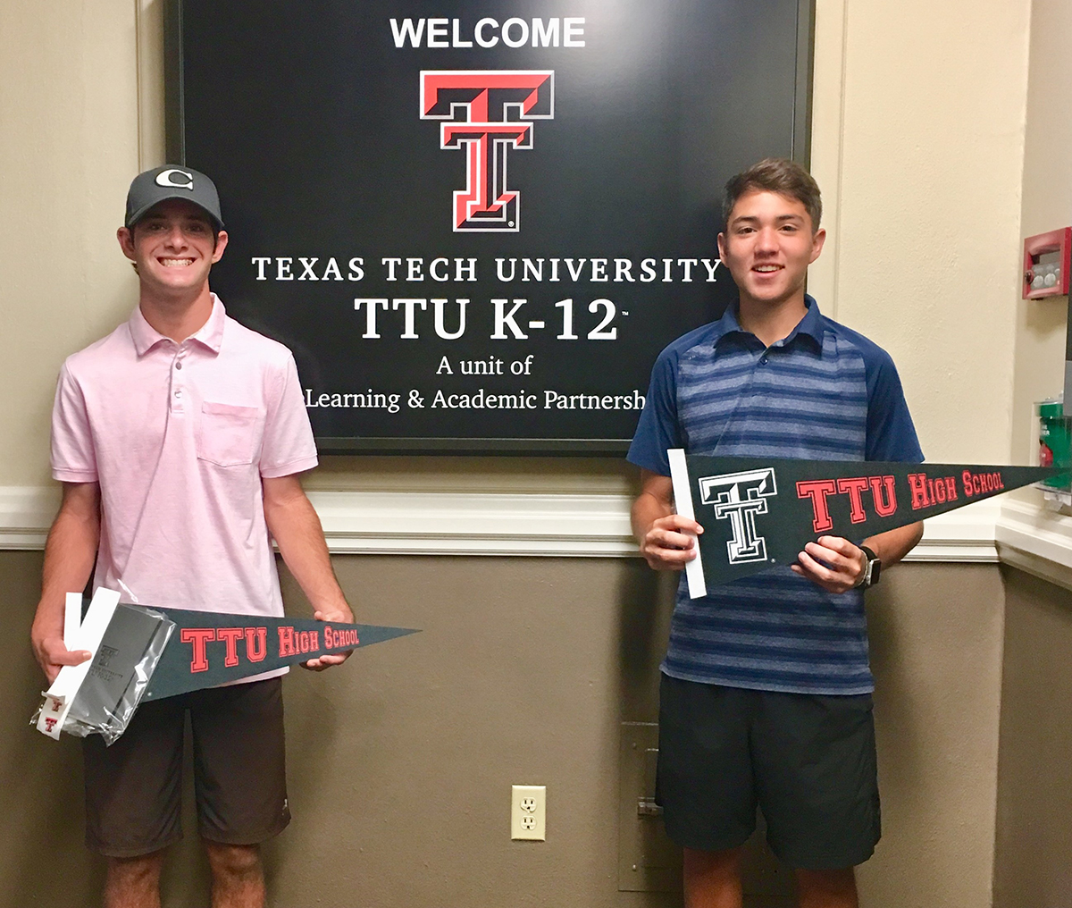 Mikey (left) and Zach drop in to visit the TTU K-12 administrative offices. Mikey and Zach stand next to one another one both sides of a TTU K-12 framed image hanging from the wall as they both hold TTU K-12 pennant flags