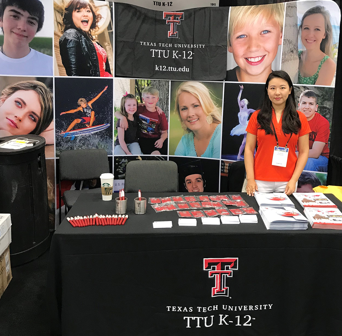 A young woman wearing a red texas tech shirt stands behind a table with ttu k-12 merchandise spread across while also standing in front of a large TTU K-12 banner