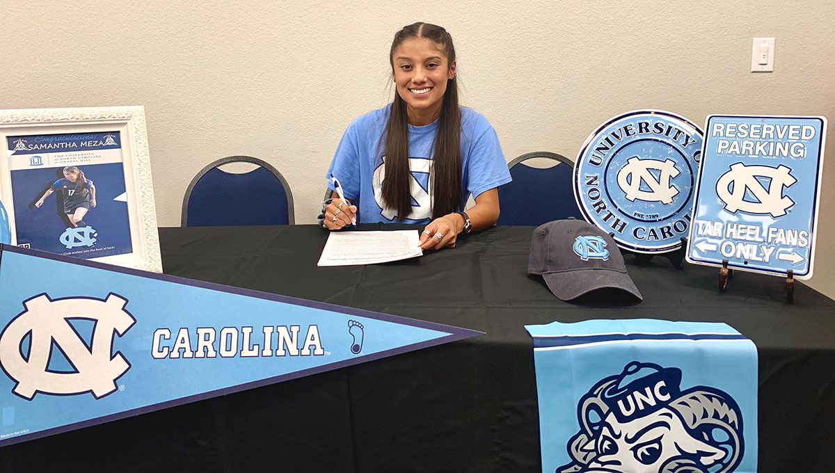 Samantha sits behind a rectangular table with university of North Carolina merchandise resting on the table as she smiles and holds a pen in her right hand papers in her left