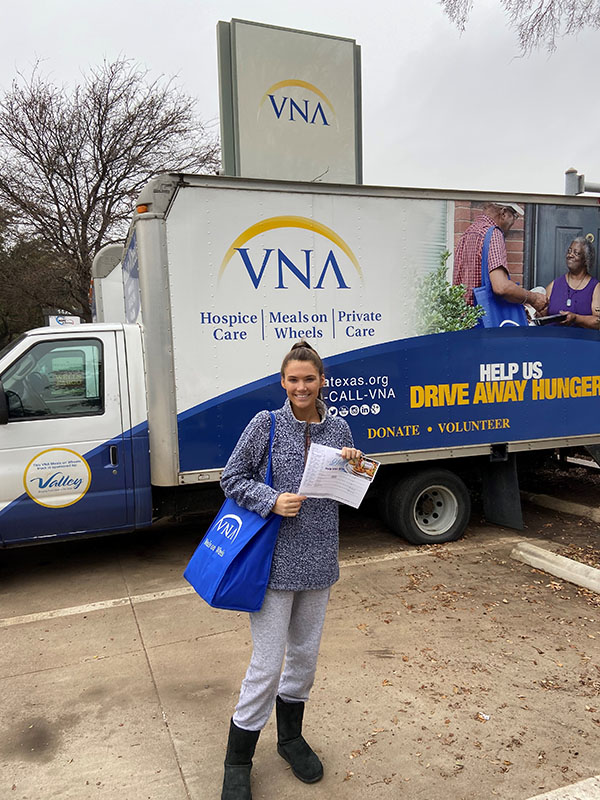A young woman stands outside with a blue bag and a paper in her hand while in front of a meals on wheels van