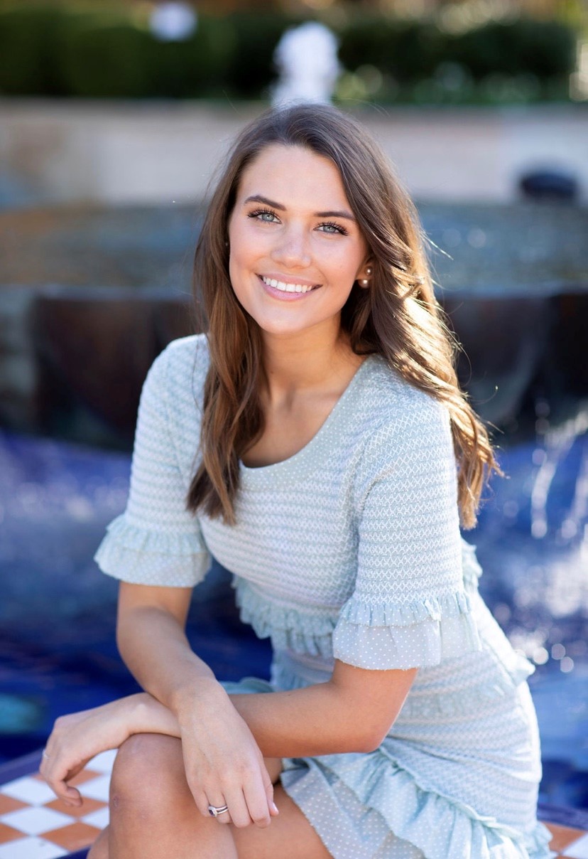 A young woman with brunette hair and a light blue colored dress sits outdoors while posing and smiling at the camera