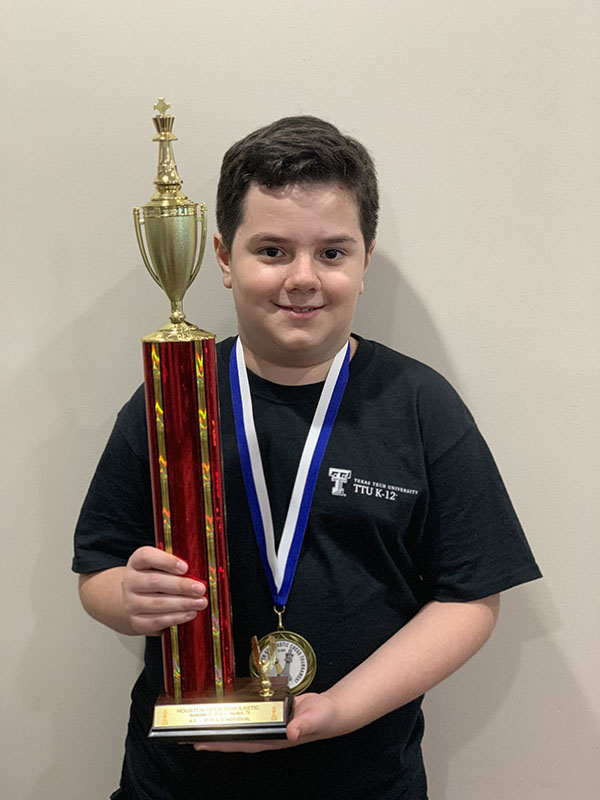 A young boy wears a medal around his neck and holds a trophy with red and yellow streaks in his hands while smiling at the camera wearing a ttu k-12 shirt