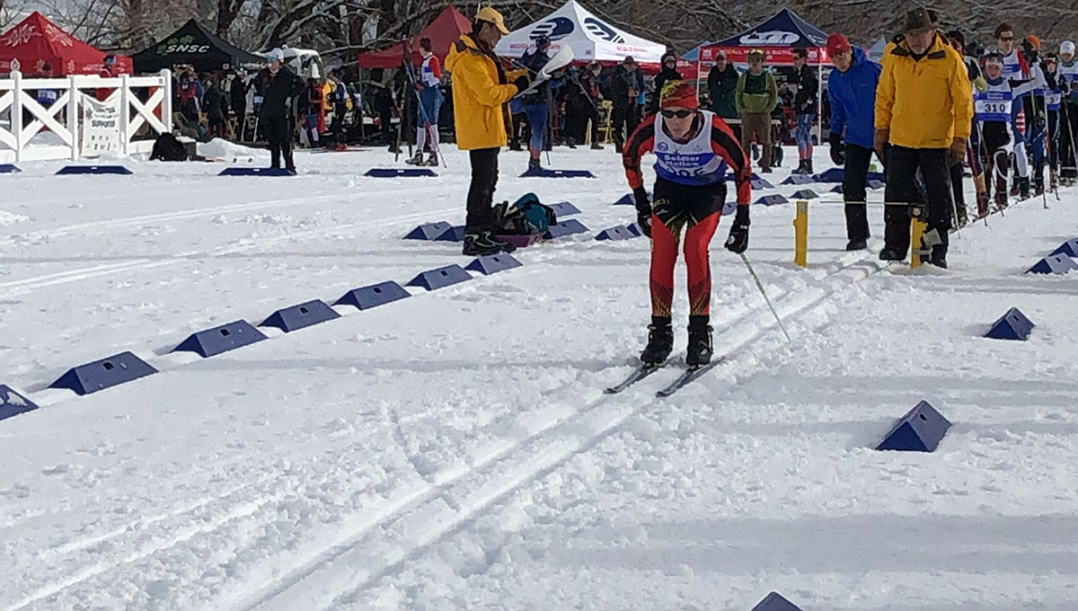 A young man wearing ski gear crouches slightly in his skis with poles in his hands on a snowy terrain with onlookers behind him.