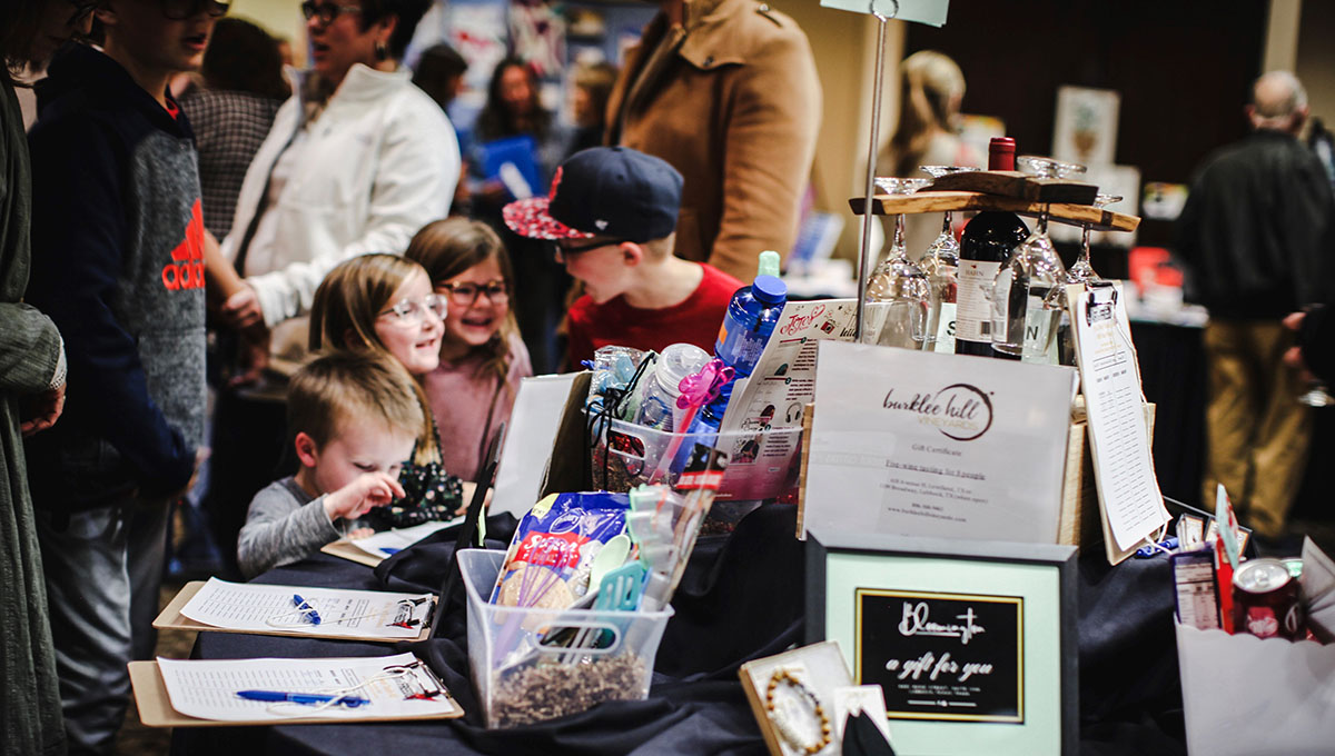 The idea for OneVoiceHome’s gala, The Art of Freedom, was conceived by teenager Garland Spore. Three small children stand among other adults as they converse and observe a table setting with a wine bottle and hold holder along with numerous other items
