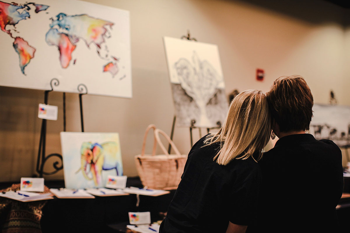 A woman with blond hair leans her head against the head of another woman as both women observe various artworks