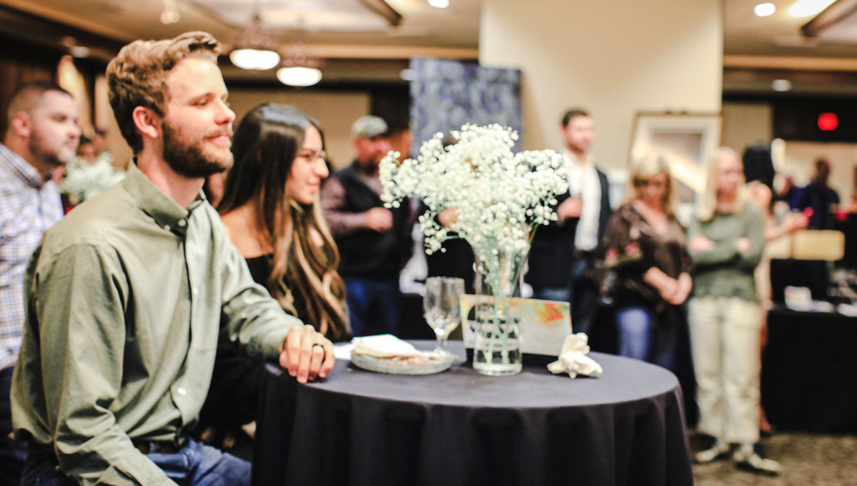 About 500 people attended The Art of Freedom, which raised funds for OneVoiceHome. A man wearing a lime green colored shirt sits at a round table with a black draping and centerpiece and looks offscreen with his left arm resting on the tale top as other onlookers in the background stand