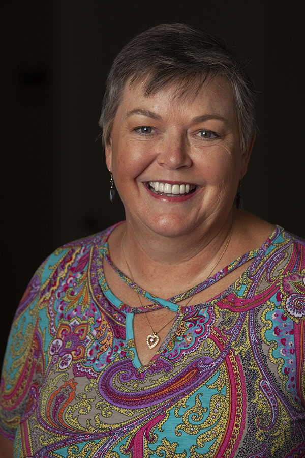 A headshot of a woman wearing a heart shaped necklace and multicolored shirt as she smiles at the camera