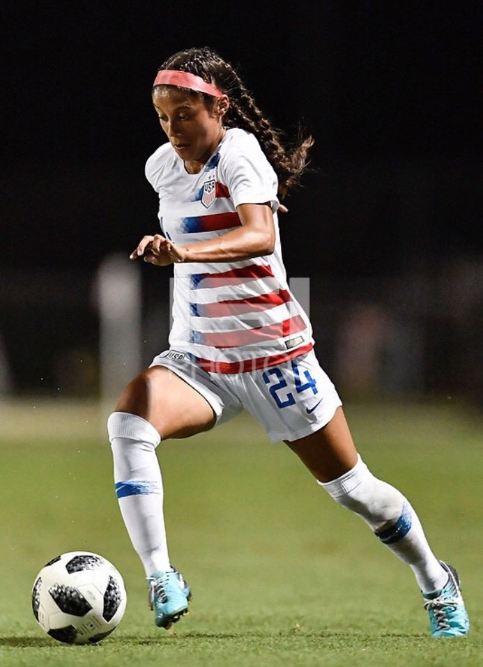 Samantha strides with her right leg ahead of her left as she prepares to kick a soccer ball while looking down at the ball