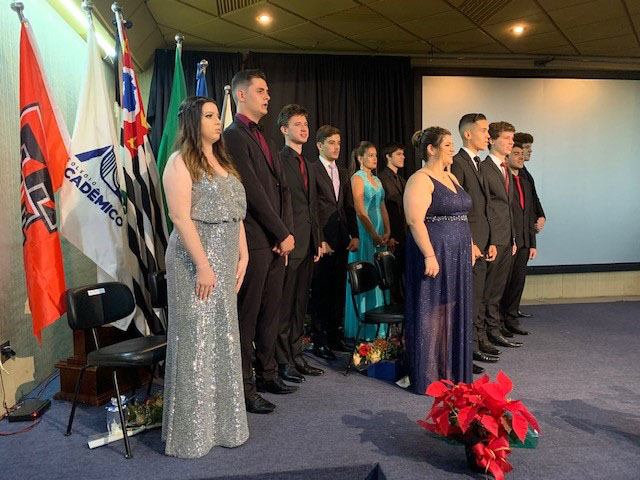 Senior and junior students stand on a stage in formal attire and look offscreen.