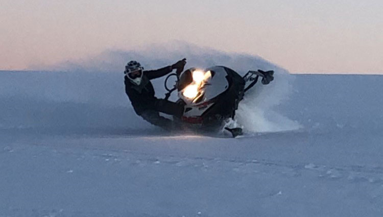 Hudson takes a wild ride on a snow mobile. A young man on a snowmobile drifts the snowmobile slightly as it glides across a snowy landscape.
