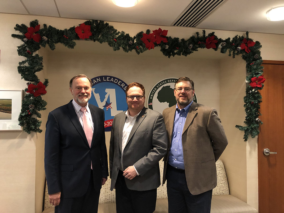 Three men stand side by side indoors in front of an wall with a wreath over the top