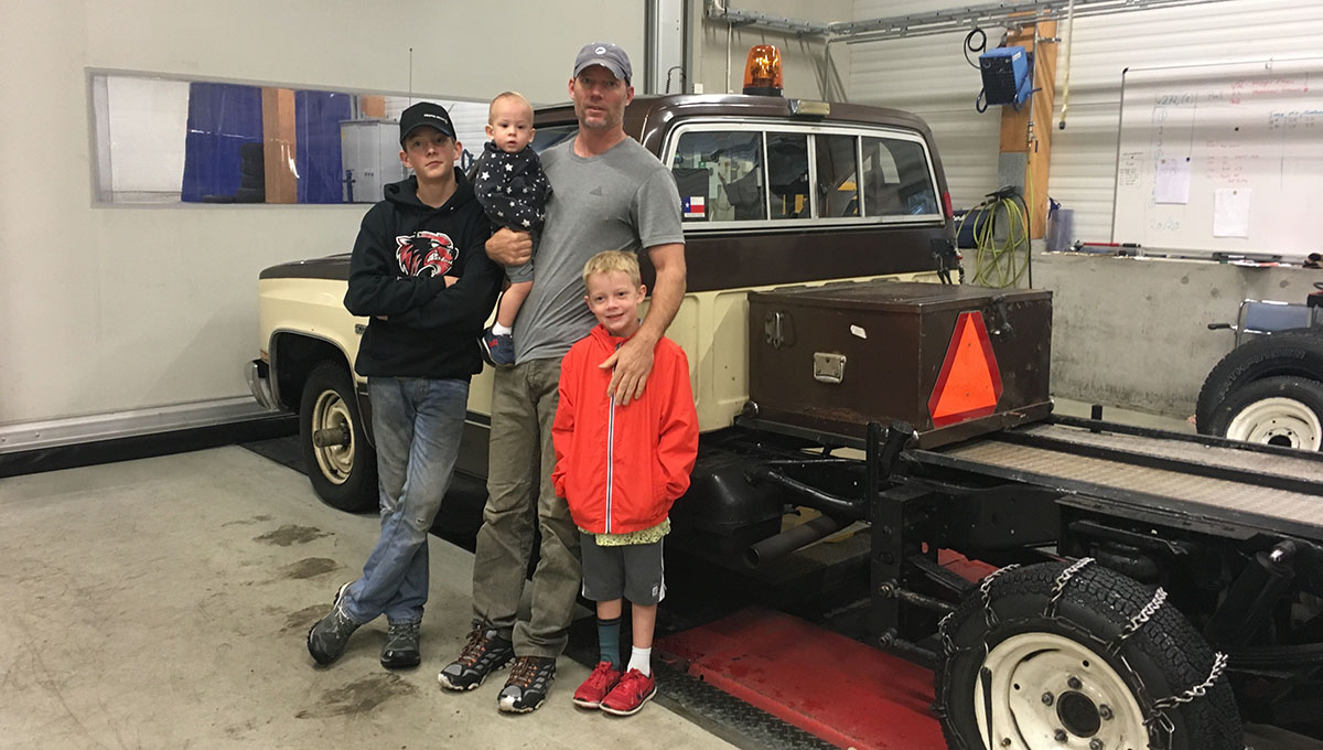 A father leans against a truck with snow tires as he holds a baby boy in his right arm and rests his left arm over the chest of his young son with his oldest son standing to his right with his arms crossed.