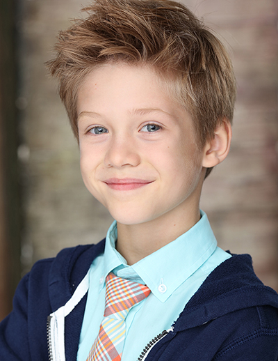 A young boy wearing a teal colored shirt and teal and orange necktie with a dark blue jacket smiles at the camera