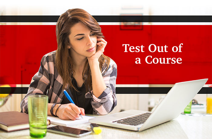 A female sits behind a desk while staring at a laptop computer and holding a blue writing utensil in her right hand with the words test out of a course in the background.