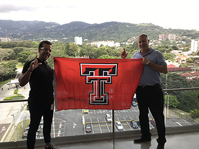 Two men hold a red and black Texas Tech University flag and stand on opposite ends of the flag on a balcony overlooking scenic Costa Rica