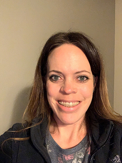 A woman with brunette hair wearing a black the north face jacket smiles at the camera while standing indoors