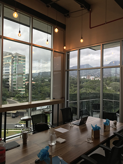 A small room is filled with a long brown table with gift bags on top along with cups, a laptop, and two beverages with chairs pushed in behind the table and a view of local greenery through the window.