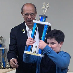 A man wearing glasses stands next to a young boy wearing a blue jacket and black shirt while he holds up a large trophy.