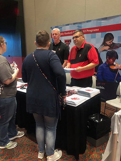 Jeff Oldham and Israel Galvan chat with attendees at the Texas School Counselor Association PSCC in San Antonio. Two men stand next to one another behind a table and face two women who stand on the opposite side of the table.