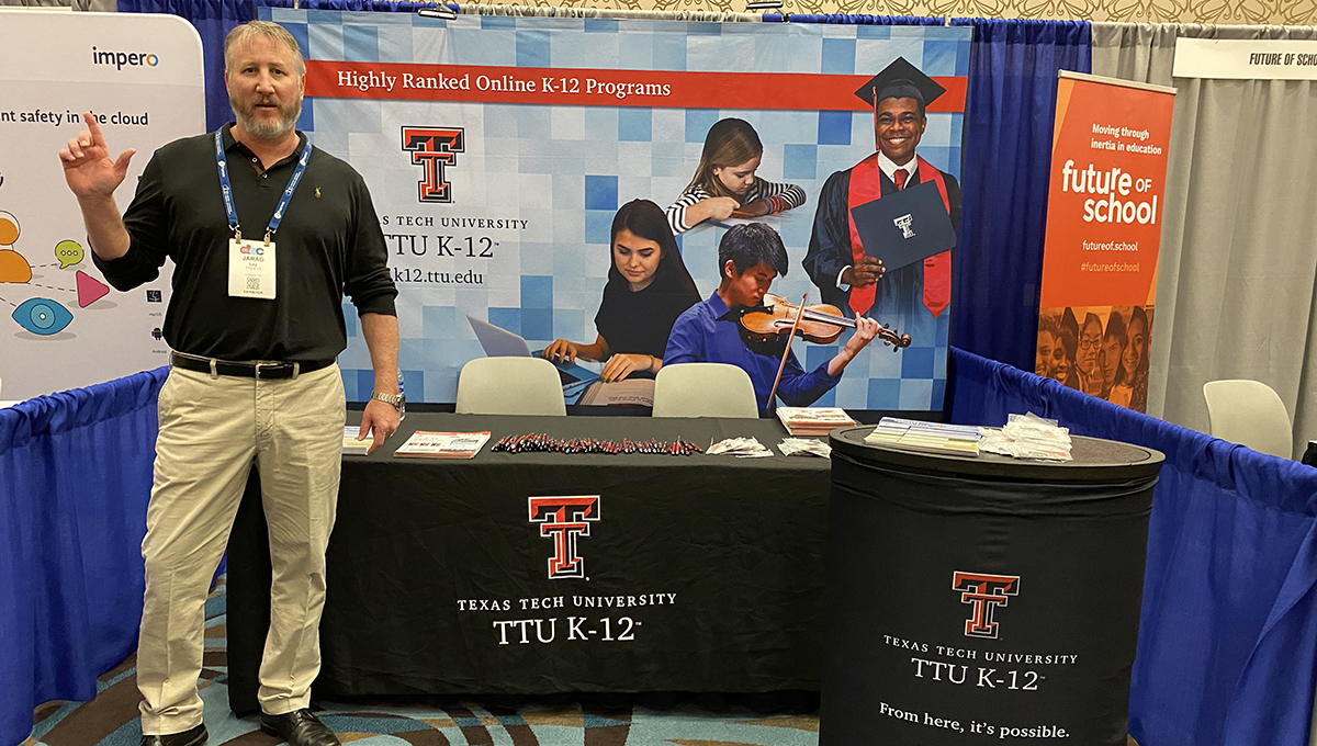 Jared Lay represents TTU K-12 at the DLAC booth in Austin. A man stands to the left of the frame performing the guns up symbol while standing in front of a TTU K-12 table and backdrop.
