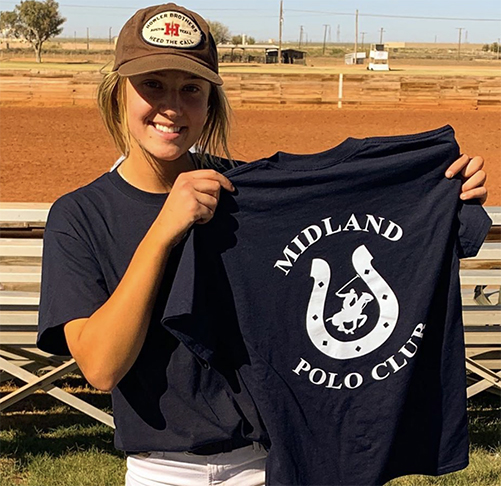  A young girl stands outdoors wearing a brown billed cap, navy blue shirt, and white pants while smiling and holding a shirt in her hands that reads Midland Polo Club