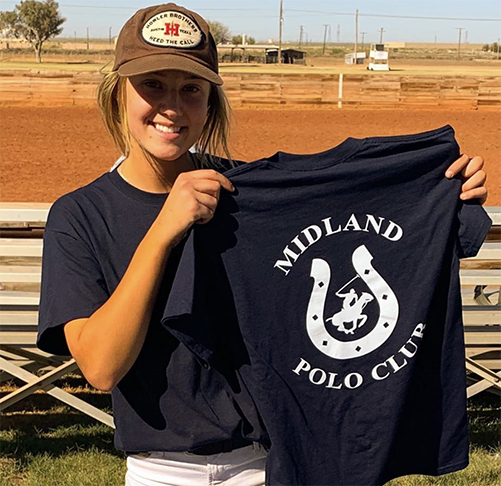 Sara is a member of the Midland Polo Club. A young girl stands outdoors wearing a brown billed cap, navy blue shirt, and white pants while smiling and holding a shirt in her hands that reads Midland Polo Club