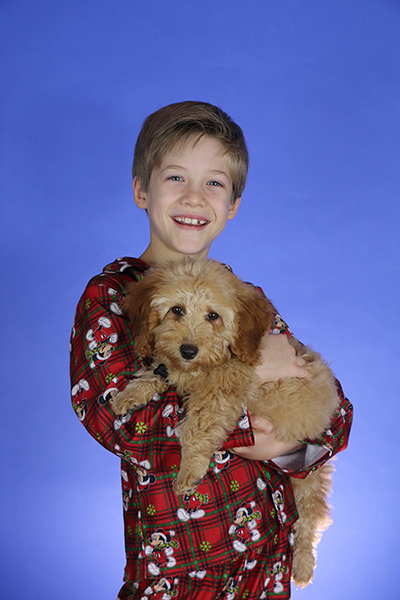 A young boy wearing red pajamas with mickey mouse print holds a brown colored dog in his arms and smiles while standing in front of a blue background