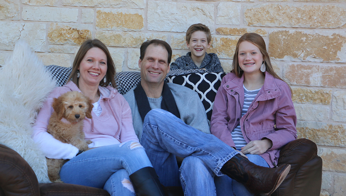 River with his family and Gypsy. A man, woman, and their teenage daughter sit on a brown couch smiling while the woman holds a small dog in her arm and a young boy stands behind the couch while smiling