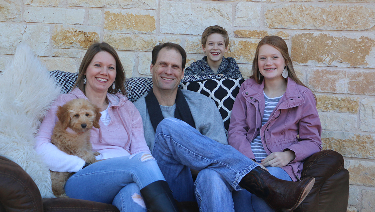 A man, woman, and their teenage daughter sit on a brown couch smiling while the woman holds a small dog in her arm and a young boy stands behind the couch while smiling 