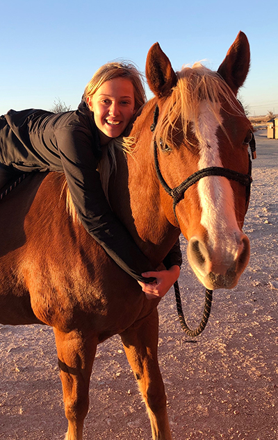 Sarah poses with her polo partner, Sysco A young girl lies on her stomach while on the back of a brown horse while outdoors