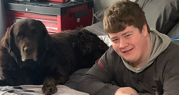 A young man rests on his stomach atop a bed next to a dark brown dog.