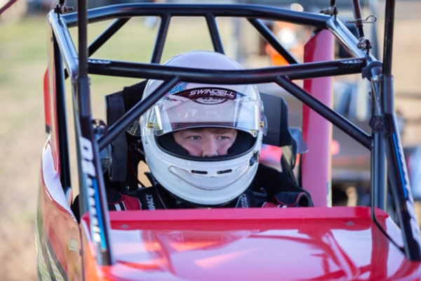 A young man wearing a white helmet stares intensely as he sits in a red race car.